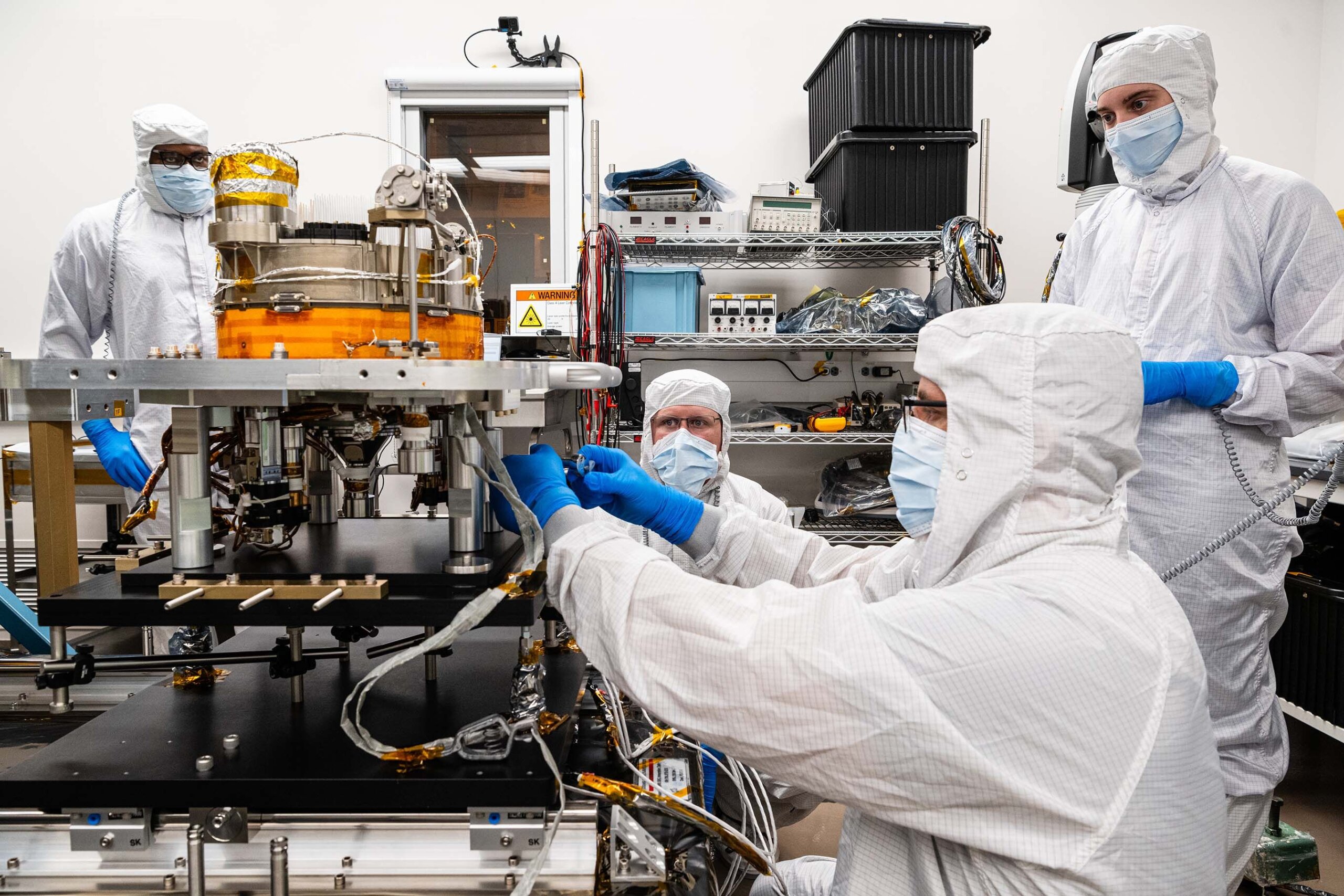 scientist bent down in white suit and gloves works on space technology in a lab with other scientists standing by